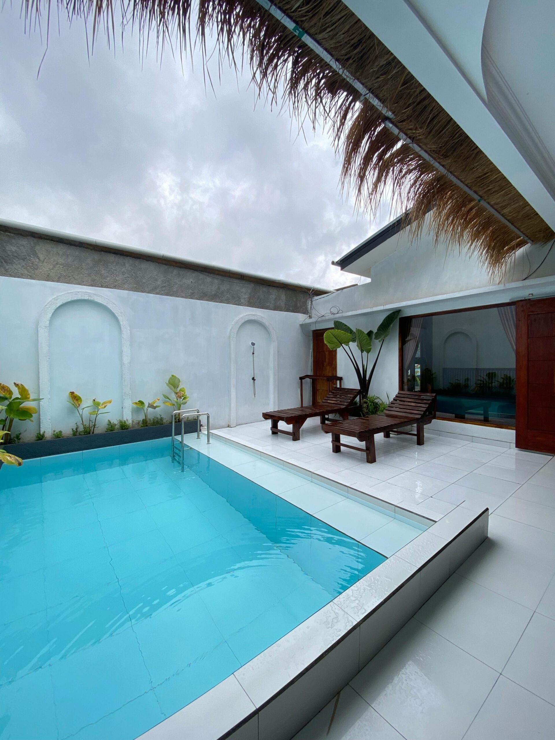 Spacious modern patio with pool, wooden loungers, and tropical plants under a cloudy sky.